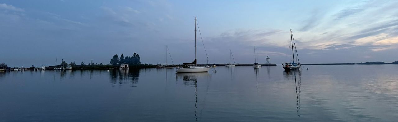 Sailboats moored on calm water at dusk under a pale moon, with a low breakwater and a small white light tower in the distance
