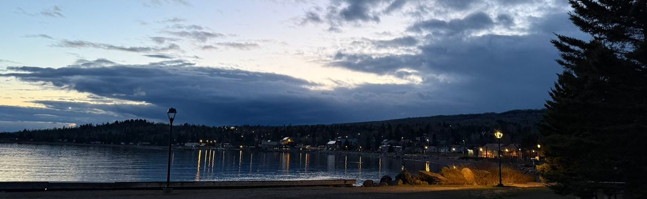 House and street lights reflecting on a harbor at dusk, with hills behind the town