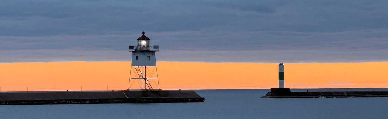 A white skeleton lighthouse with its lamp lit on the end of a breakwater, under dark cloud and an orange horizon