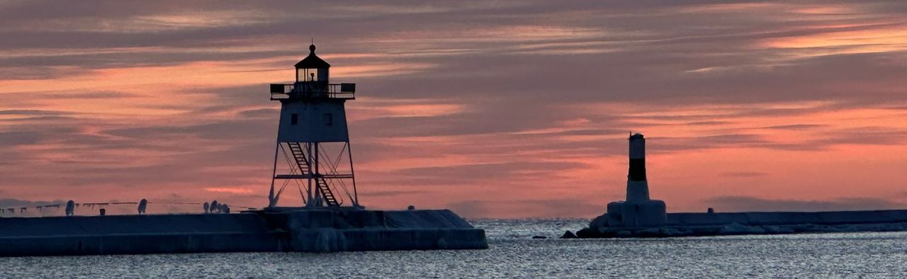 A skeleton-frame light tower on the end of a breakwater and a smaller banded light beyond it, in silhouette against a pink and orange sky