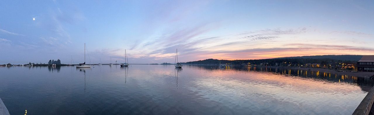 A wide panorama of a calm harbor at sunset, with sailboats at their moorings, the moon overhead and the lights of a town along the right-hand shore