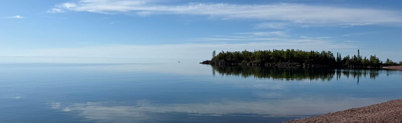 A wooded, rocky point reflected in calm blue water under thin clouds, with a red gravel beach in the foreground
