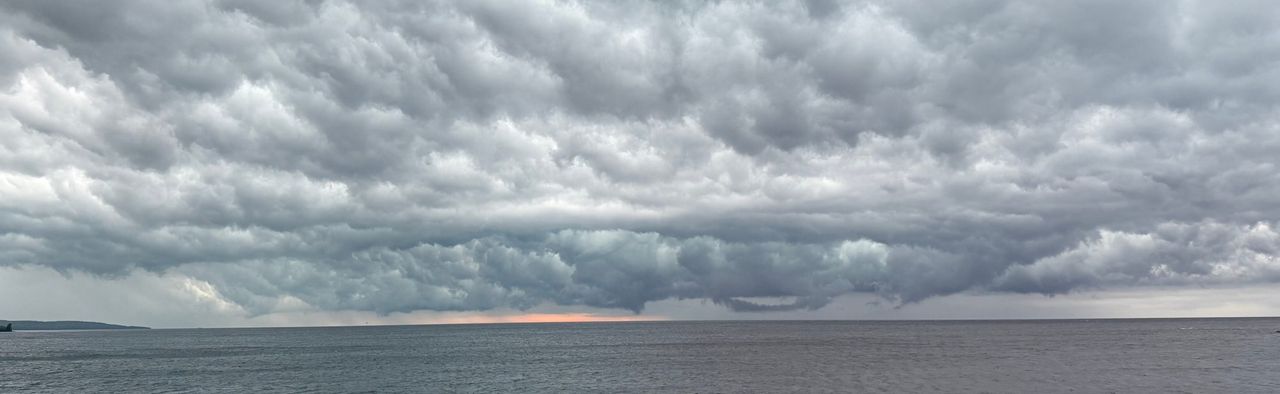 A heavy shelf of gray storm cloud over open water, with a thin orange band at the horizon, seen from a gravel shore over a green shrub