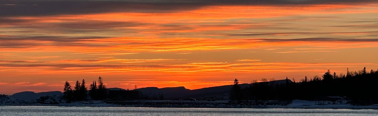 Layered ridges and a wooded, snowy shoreline in silhouette under bands of orange sunset cloud, seen across open water